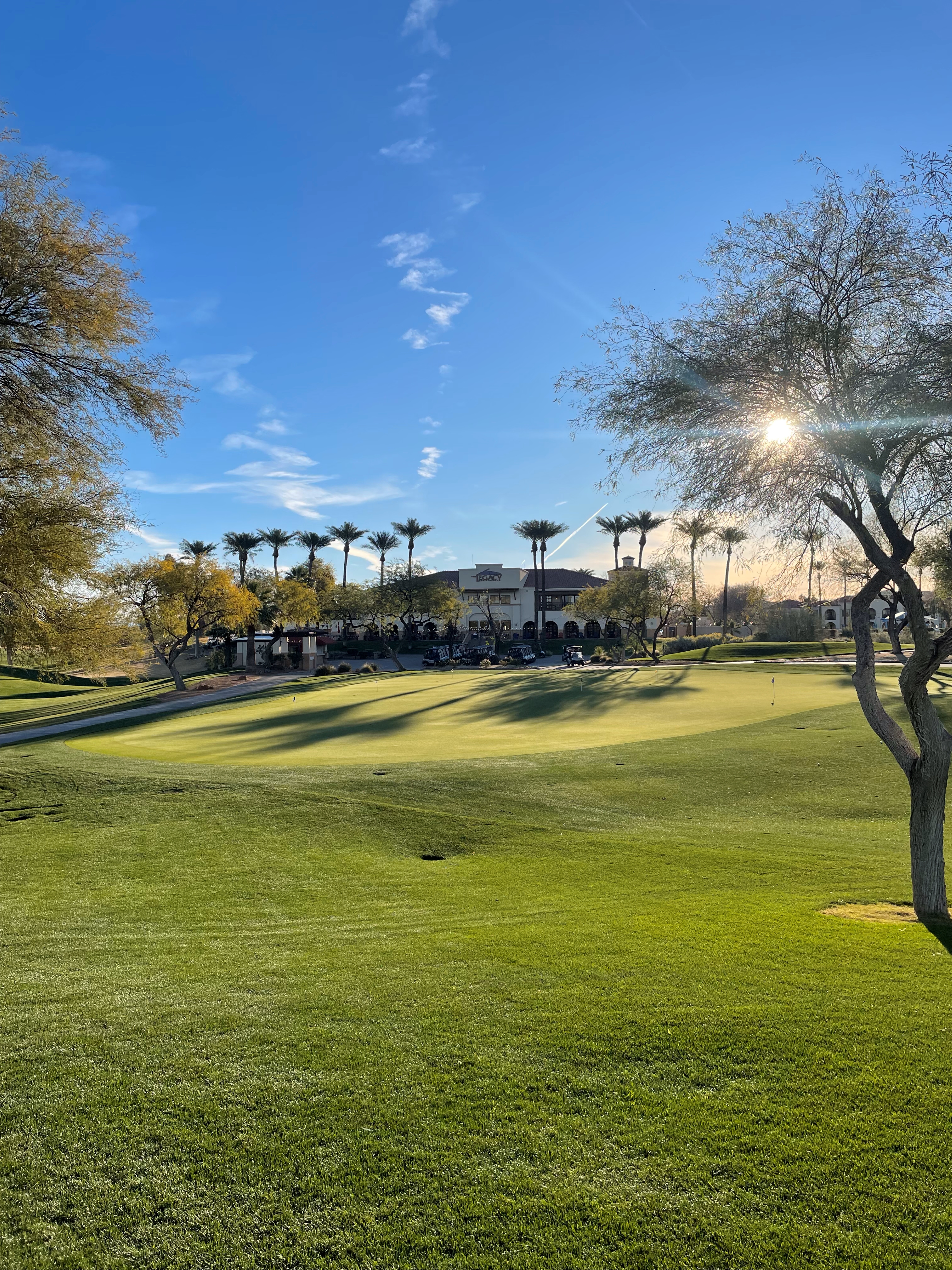 A beautifully lit practice putting green at golden hour with the Legacy Golf Club clubhouse featuring palm trees and Spanish-style architecture visible in the background under a vibrant blue sky.